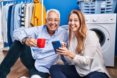 Orta yaşlı erkek ve kadın çift kahve içip akıllı telefon kullanarak çamaşır odasında çamaşır makinesini bekliyor.