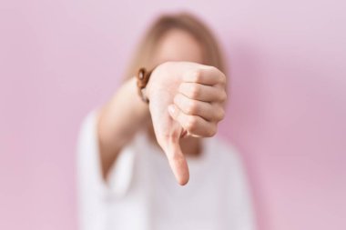 Young caucasian woman standing over pink background looking unhappy and angry showing rejection and negative with thumbs down gesture. bad expression. 