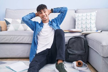 Genç İspanyol bir adam yerde oturmuş üniversite için çalışıyor rahatlıyor ve esniyor, kolları ve elleri başının arkasında mutlu bir şekilde gülümsüyor. 