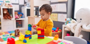 Adorable hispanic boy playing with construction blocks standing at kindergarten