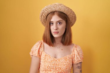 Sarı arka planda duran genç kızıl saçlı kadın. Yazlık şapka takıyor. Yüzünde ciddi bir ifade var. Basit ve doğal bir şekilde kameraya bakıyorsun.. 