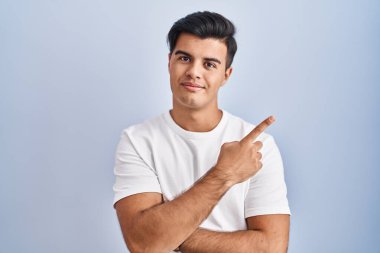 Hispanic man standing over blue background pointing with hand finger to the side showing advertisement, serious and calm face 