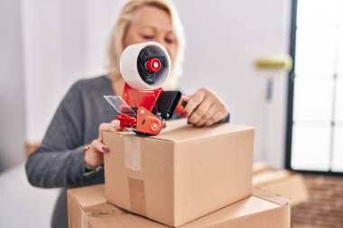 Middle age blonde woman smiling confident packing cardboard box at new home