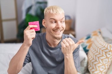 Kafkasyalı genç bir adam elinde prezervatifle yatağın kenarına oturmuş, ağzı açık bir şekilde gülümsüyor. 