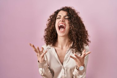 Hispanic woman with curly hair standing over pink background crazy and mad shouting and yelling with aggressive expression and arms raised. frustration concept. 