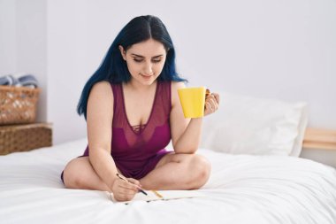 Young caucasian woman drinking cup of coffee writing on notebook at bedroom