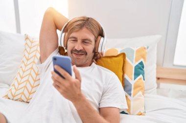 Sarışın genç adam yatakta uzanmış akıllı telefondan video izliyor.