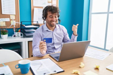 Ofiste çalışan İspanyol asıllı genç adam online alışveriş yapıyor başparmağıyla yan tarafa işaret ediyor ve ağzı açık gülümsüyor. 