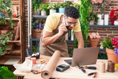 Young arab man florist talking on smartphone using laptop at florist