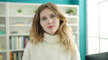 Young blonde woman student standing with relaxed expression at library university