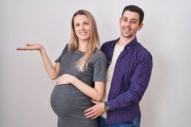 Genç bir çift beyaz arka planda duran bir bebek bekliyor. Gülümseyen, mutlu bir şekilde sunan ve avuç içi kameraya bakan bir çift.. 