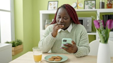 Örgülü saçlı Afrikalı kadın yemek odasında akıllı telefon kullanarak kurabiye yiyor.