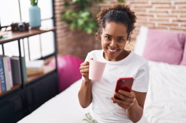 African american woman using smartphone drinking coffee at bedroom