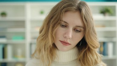 Young blonde woman student standing with relaxed expression at library university