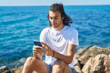 Young hispanic man using smartphone sitting on rock at seaside