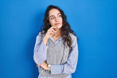 Mavi arka planda duran genç esmer kadın, elleri çenesinde soru sorarken, düşünceli bir yüz ifadesi vardı. Düşünceli bir yüzle gülümsüyor. şüphe kavramı. 