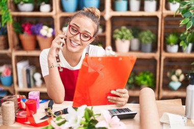 Young beautiful hispanic woman florist talking on smartphone reading clipboard at florist