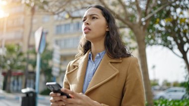 Genç, güzel, İspanyol bir kadın parkta ciddi yüzlü akıllı telefon kullanıyor.