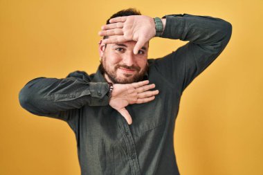 Plus size hispanic man with beard standing over yellow background smiling cheerful playing peek a boo with hands showing face. surprised and exited 