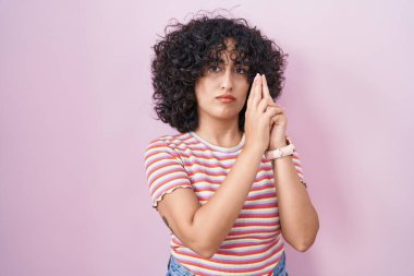 Orta Doğulu genç bir kadın pembe arka planda duruyor. Elinde el hareketleriyle sembolik bir silah tutuyor. Cinayet silahlarıyla oynuyor. 