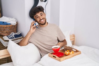 Genç Arap adam akıllı telefondan konuşuyor. Yatak odasında kahvaltı yapıyor.