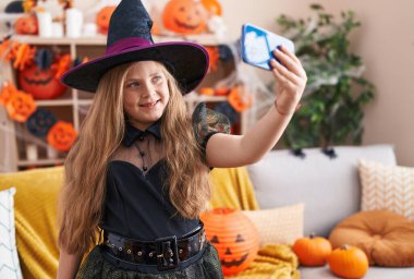 Cadı kostümü giyen sevimli sarışın kız evde akıllı telefondan selfie çekiyor.