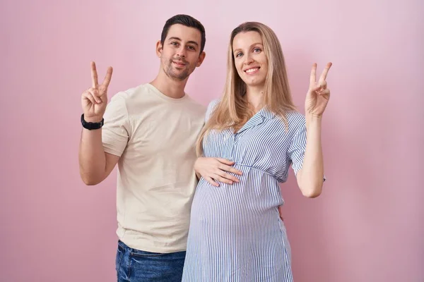 年轻夫妇期待着一个婴儿站在粉红的背景上 微笑着看着摄像机 手指头在做胜利的标志 第二点 — 图库照片
