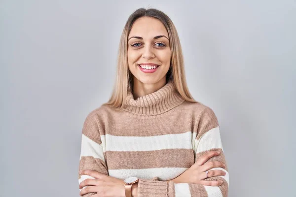 Young blonde woman wearing turtleneck sweater over isolated background happy face smiling with crossed arms looking at the camera. positive person.