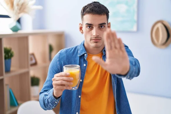 Handsome hispanic man drinking glass of orange juice with open hand doing stop sign with serious and confident expression, defense gesture