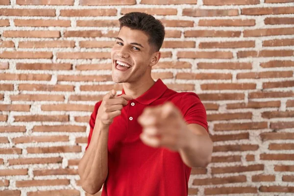 若いヒスパニック系の男がレンガの壁の上に幸せと面白い顔を持つカメラに指を指して立っている 良いエネルギーと振動は — ストック写真
