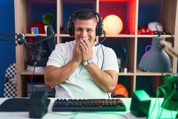 若いヒスパニック系の男がビデオゲームをして笑って恥ずかしがり屋のクスクス笑いを手で口をカバー ゴシップやスキャンダルの概念 — ストック写真