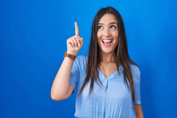 若いブルネットの女性が成功したアイデアで指を指して青い背景の上に立っています 興奮して幸せよ 第1位 — ストック写真