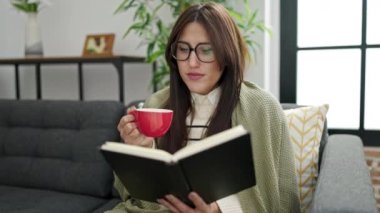 Young beautiful hispanic woman reading book and drinking coffee sitting on sofa at home