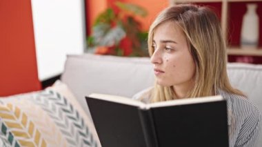 Young blonde woman reading book sitting on sofa at home