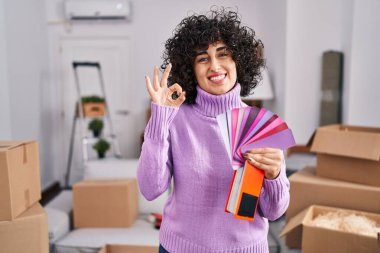 Yeni ev duvarının rengini seçen kıvırcık saçlı genç esmer kadın parmaklarıyla işaret veriyor, gülümseyerek mükemmel bir sembol oluşturuyor. 