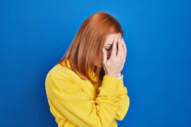 Young woman standing over blue background with sad expression covering face with hands while crying. depression concept. 