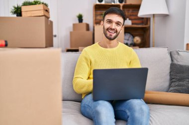 Young hispanic man using laptop sitting on sofa at new home