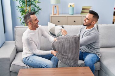 Two men couple fighting with cushion sitting on sofa at home