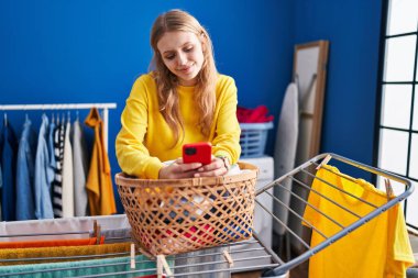 Young blonde woman using smartphone hanging clothes on clothesline at laundry room