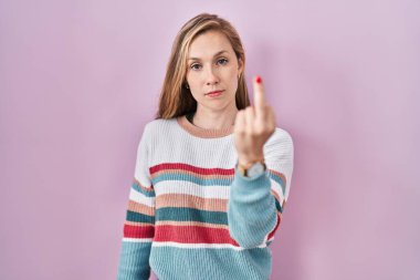 Young blonde woman standing over pink background showing middle finger, impolite and rude fuck off expression 