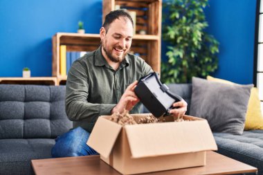 Young hispanic man unpacking virtual reality glasses sitting on sofa at home