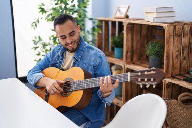 Young hispanic man playing classical guitar sitting on table at home