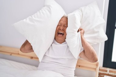 Yaşlı gri saçlı adam yatak odasındaki gürültü için kulaklarını kapatıyor.
