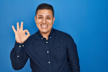 Hispanic young man standing over blue background smiling positive doing ok sign with hand and fingers. successful expression. 