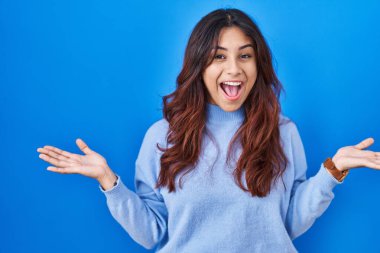 Hispanic young woman standing over blue background celebrating crazy and amazed for success with arms raised and open eyes screaming excited. winner concept 