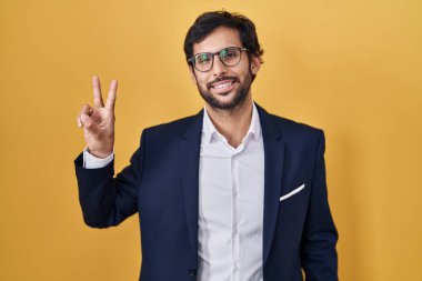 Sarı arka planda dikilen yakışıklı Latin adam kameraya bakıp zafer işareti yapıyor. İki numara.. 