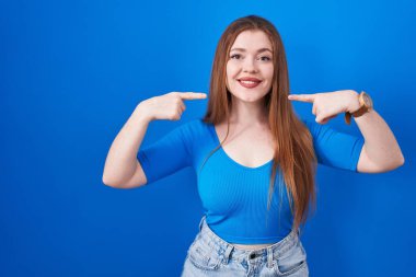 Mavi arka planda duran kızıl saçlı kadın gülümseyip, parmaklarıyla ve ağzıyla işaret ediyor. diş sağlığı kavramı. 