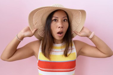 Orta yaşlı Çinli kadın pembe arka planda yaz şapkası takıyor şaşkınlık ve şaşkınlık içinde bir ifade, korku ve heyecan dolu bir yüz.. 