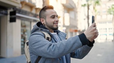 İspanyol adam sokakta akıllı telefonuyla fotoğraf çekerken kendinden emin gülümsüyor.