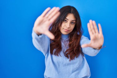 Latin kökenli genç bir kadın mavi arka planda duruyor. El ve parmakları kullanarak çerçeve yapıyor. Kamera bakış açısı. 